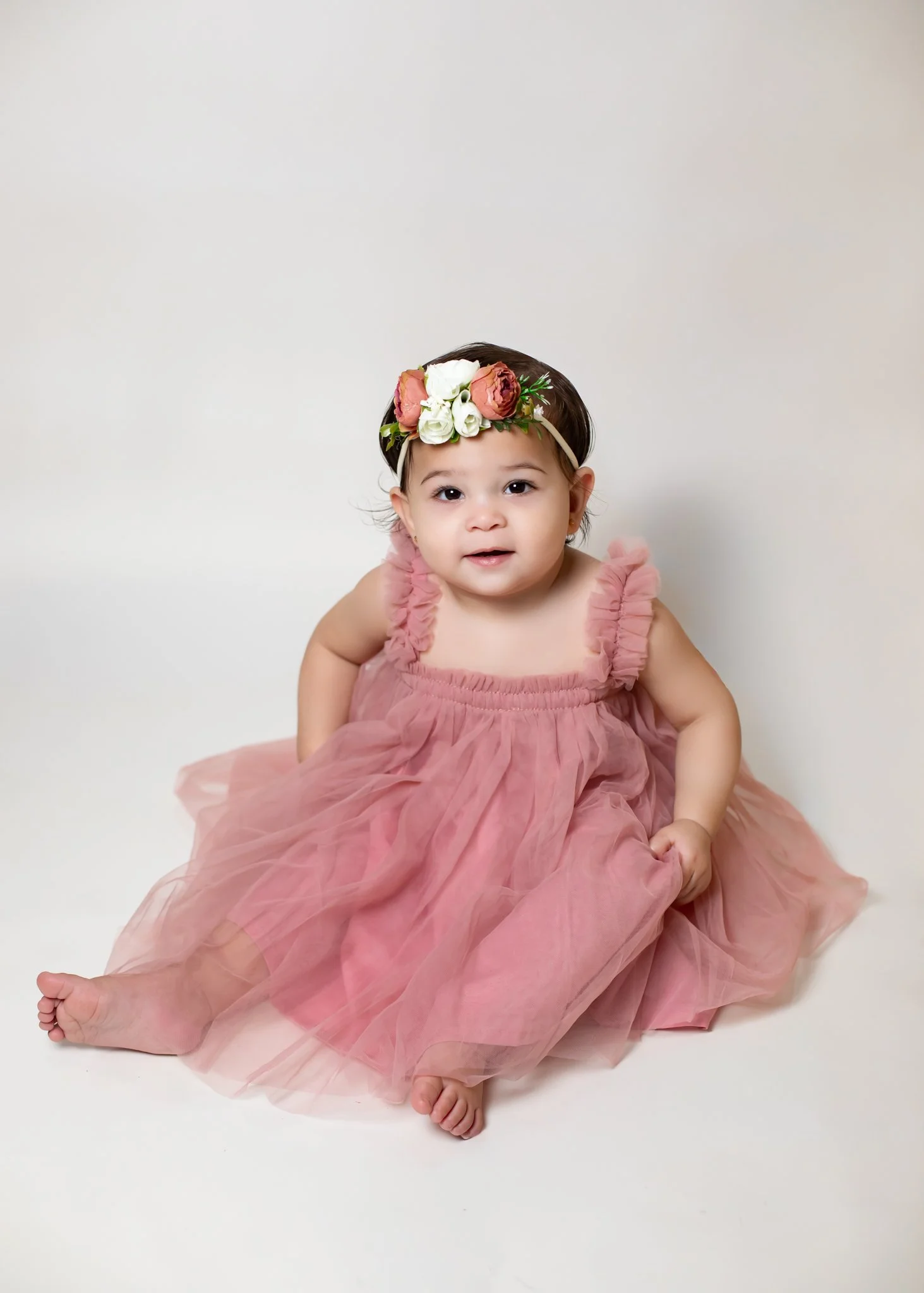 A young girl with a floral headband and a pink ruffled dress, sitting barefoot on a white background.