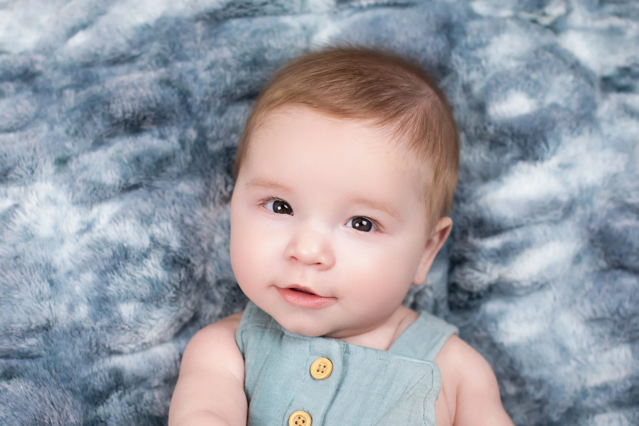 Close-up of a young child's face with short, light brown hair, lying on a soft, gray textured blanket, looking at the camera with wide eyes and a gentle expression.