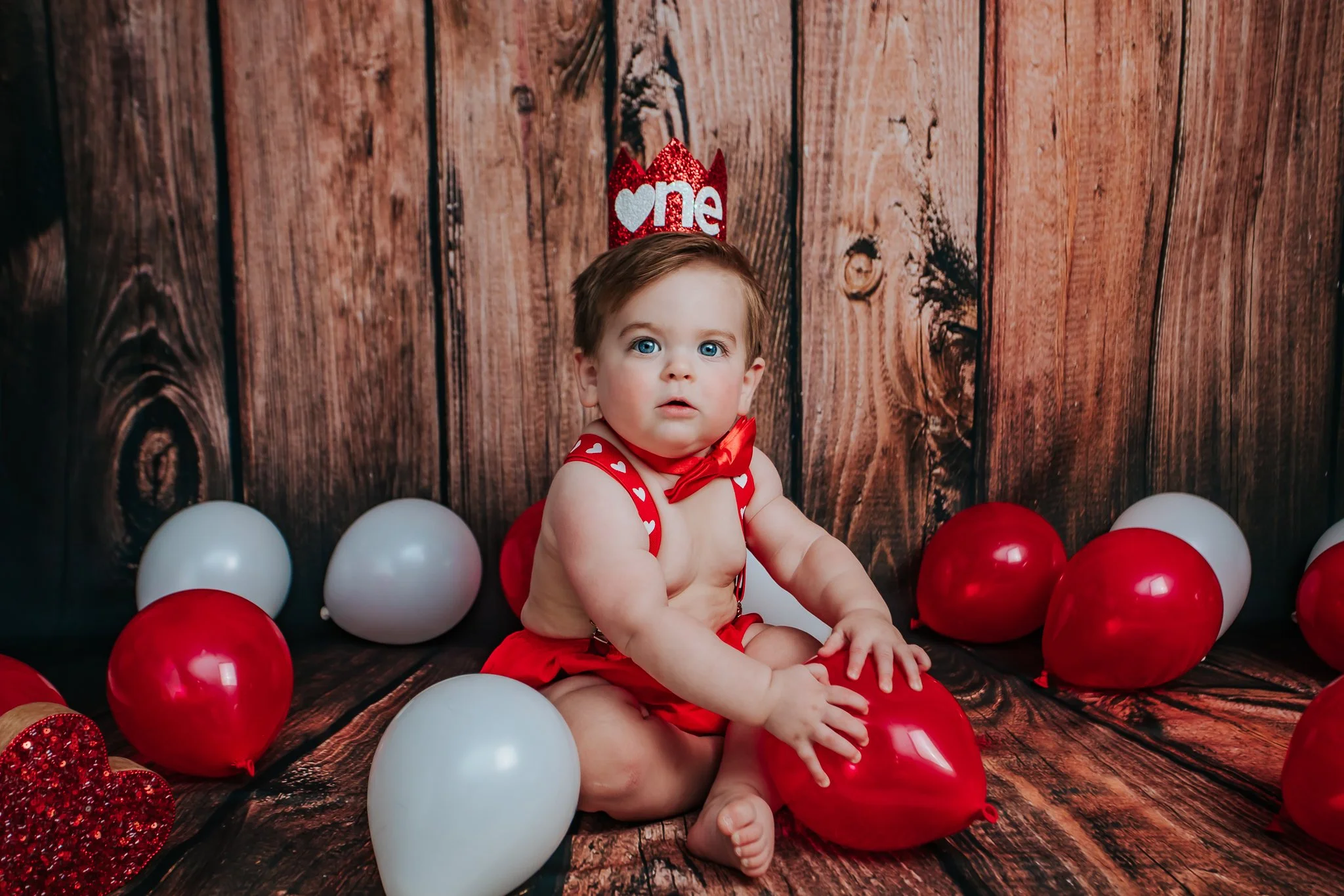 Young child with blue eyes sitting among red and white balloons, wearing a red dress with heart patterns, against a wooden background with a red sparkly crown that says 'one' on her head.