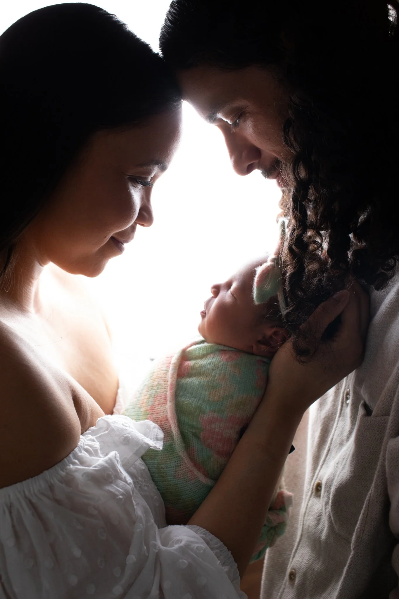 A woman, a man, and a baby are touching foreheads in a close, tender moment, backlit with soft light.