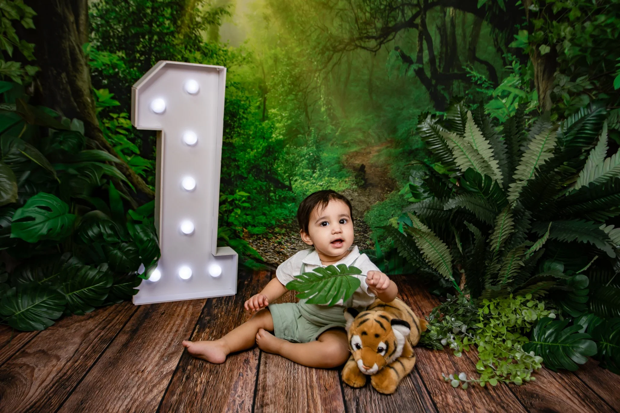 A young boy sitting on a wooden floor surrounded by lush green jungle-themed decor, including large leafy plants and a forest mural background, with a plush tiger toy in front of him.
