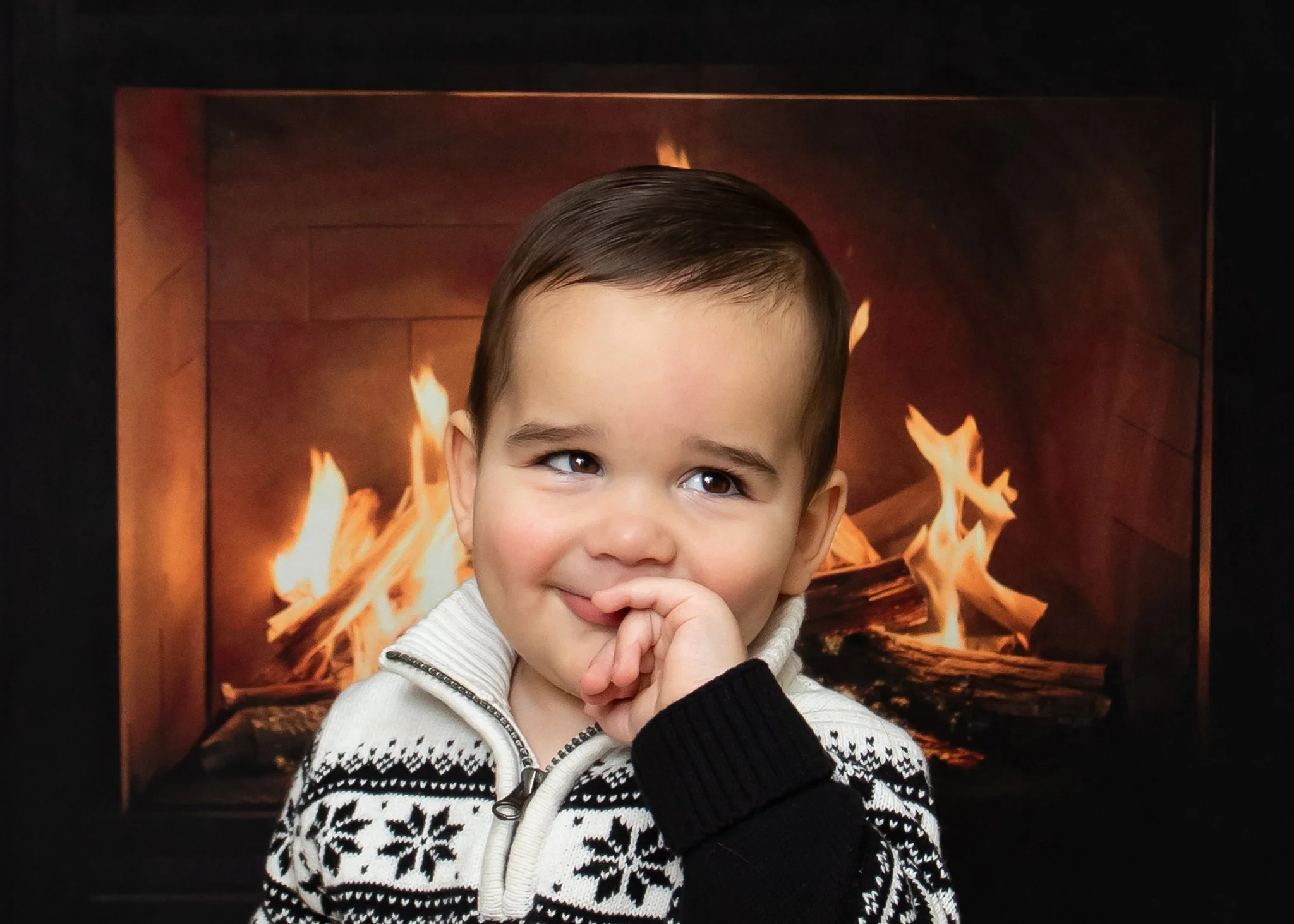 A young boy with brown hair and brown eyes is standing in front of a fireplace with flames. He is smiling slightly and has one hand near his mouth. He is wearing a white sweater with a black snowflake pattern and a zipper.