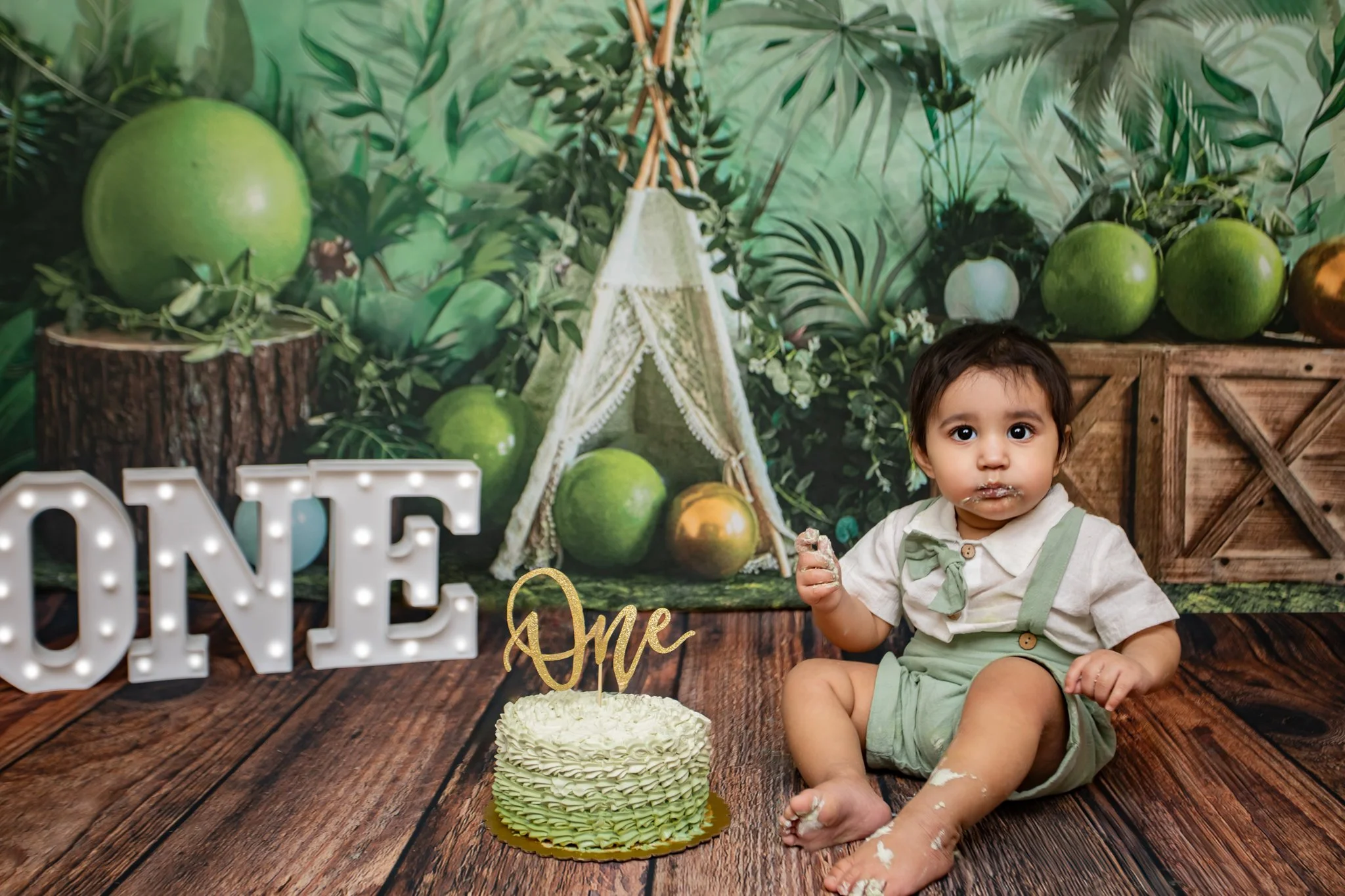 A young child with dark hair and big eyes sitting on a wooden floor, holding cake crumbs, with a green and white layered birthday cake in front of them. The background features a jungle-themed wall mural, large green fruit, and decorative items, incl