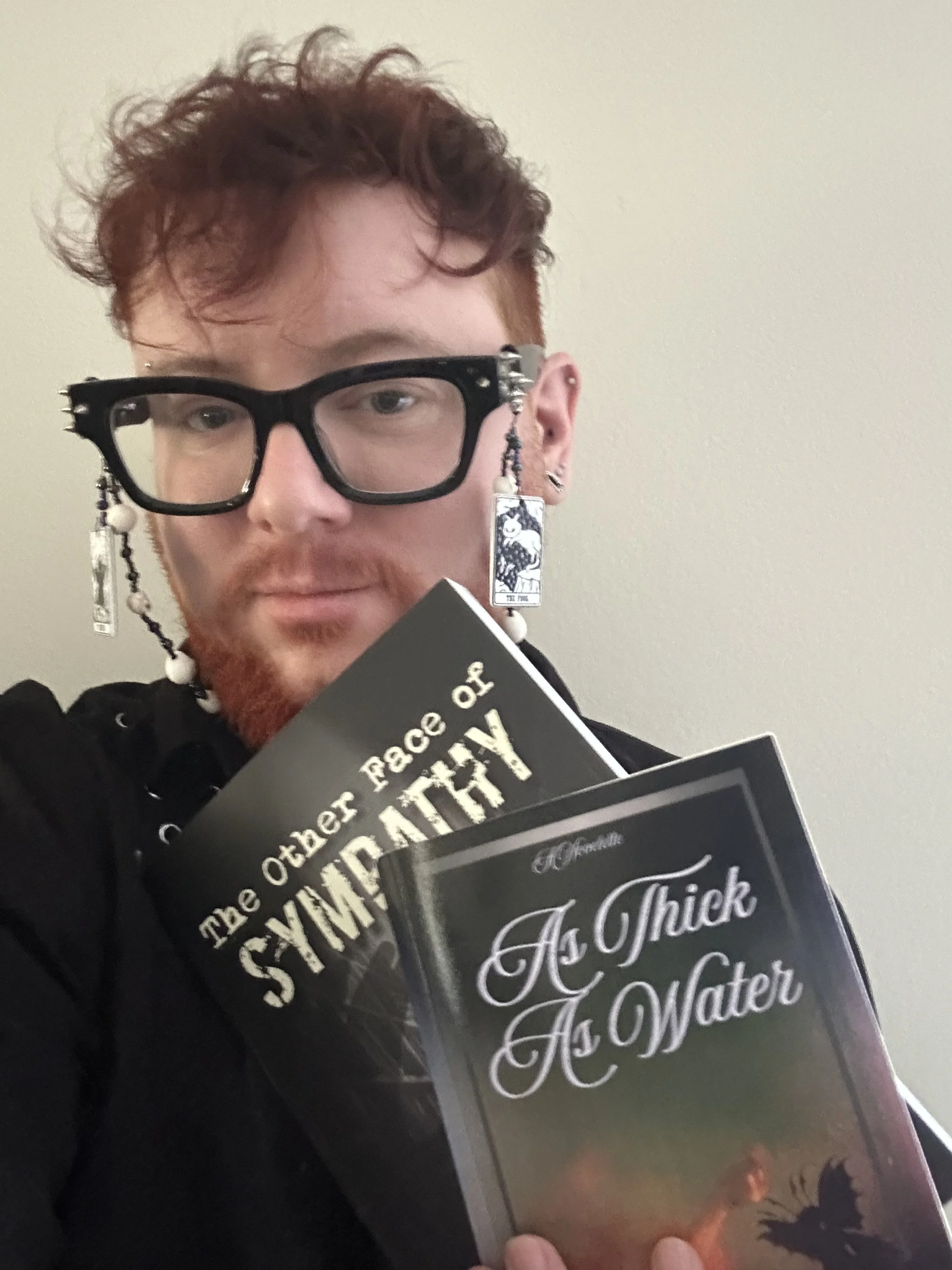 A photo of R. Haven, a man with a red beard and curly hair cropped short. He has black glasses with silver spikes on the sides, and a glasses chain with hanging tarot beads on them. He holds books, The Other Face of Sympathy and As Thick as Water.