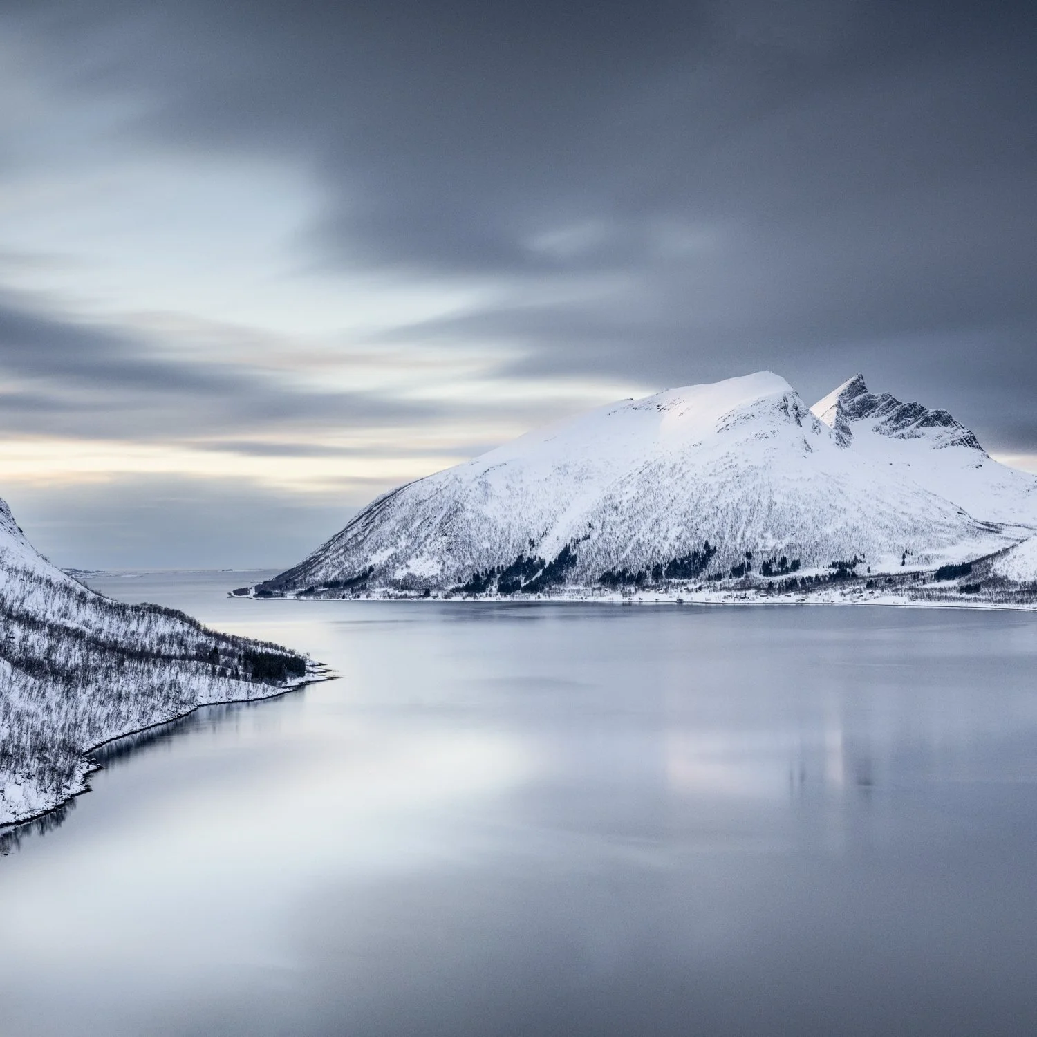 Mountain Light. Long-exposure photograph of a snow-covered mountain in Senja rising above calm water beneath a softly illuminated sky.