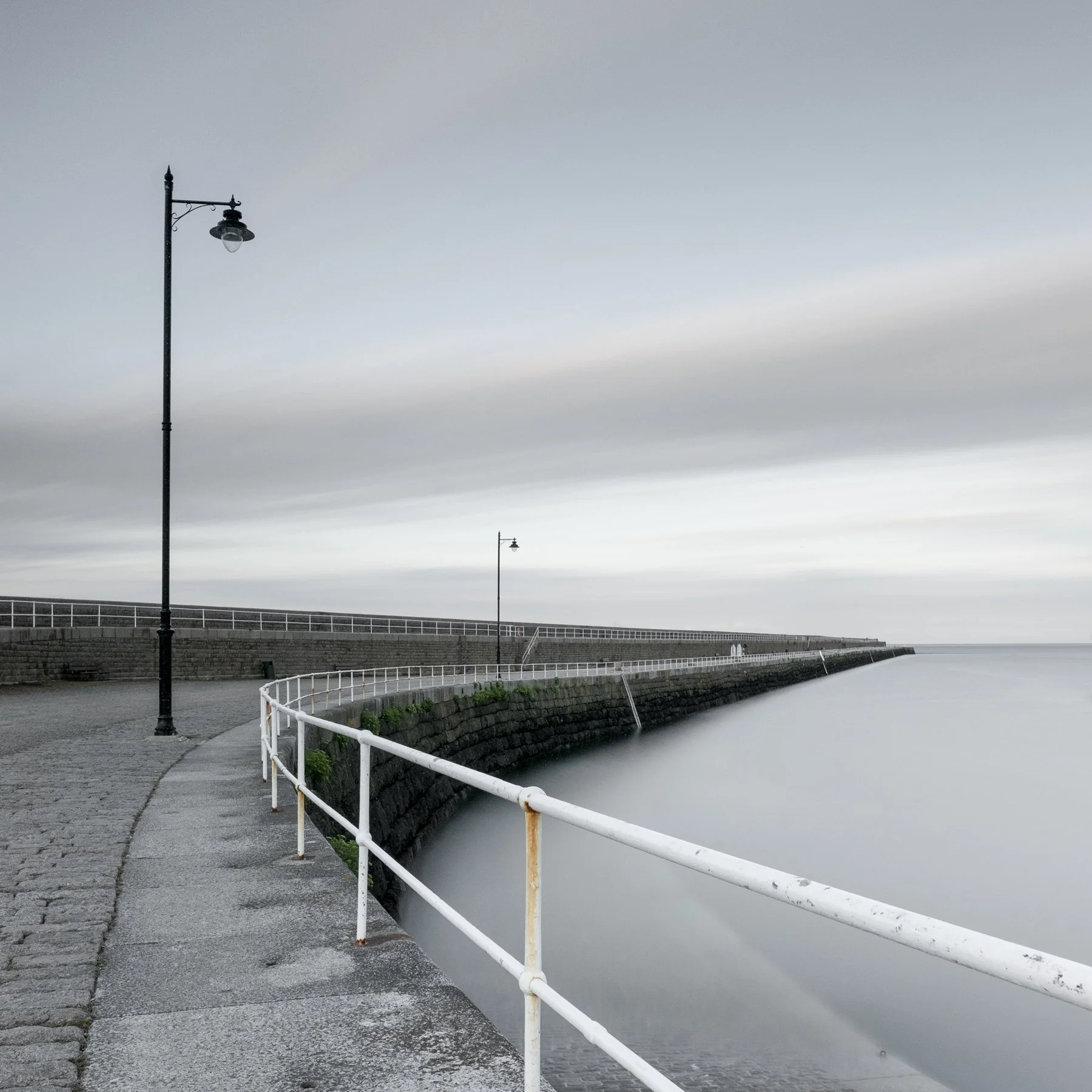 St Catherines Breakwater. Long-exposure photograph of a quiet coastal breakwater in Jersey and curved sea wall beside calm water, with a solitary lamp post.