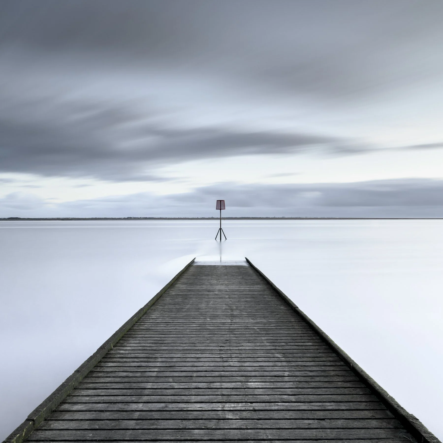 Reaching Out. Long-exposure photograph of a wooden pier extending towards a small navigation marker across calm, pale water at Lytham St Annes.