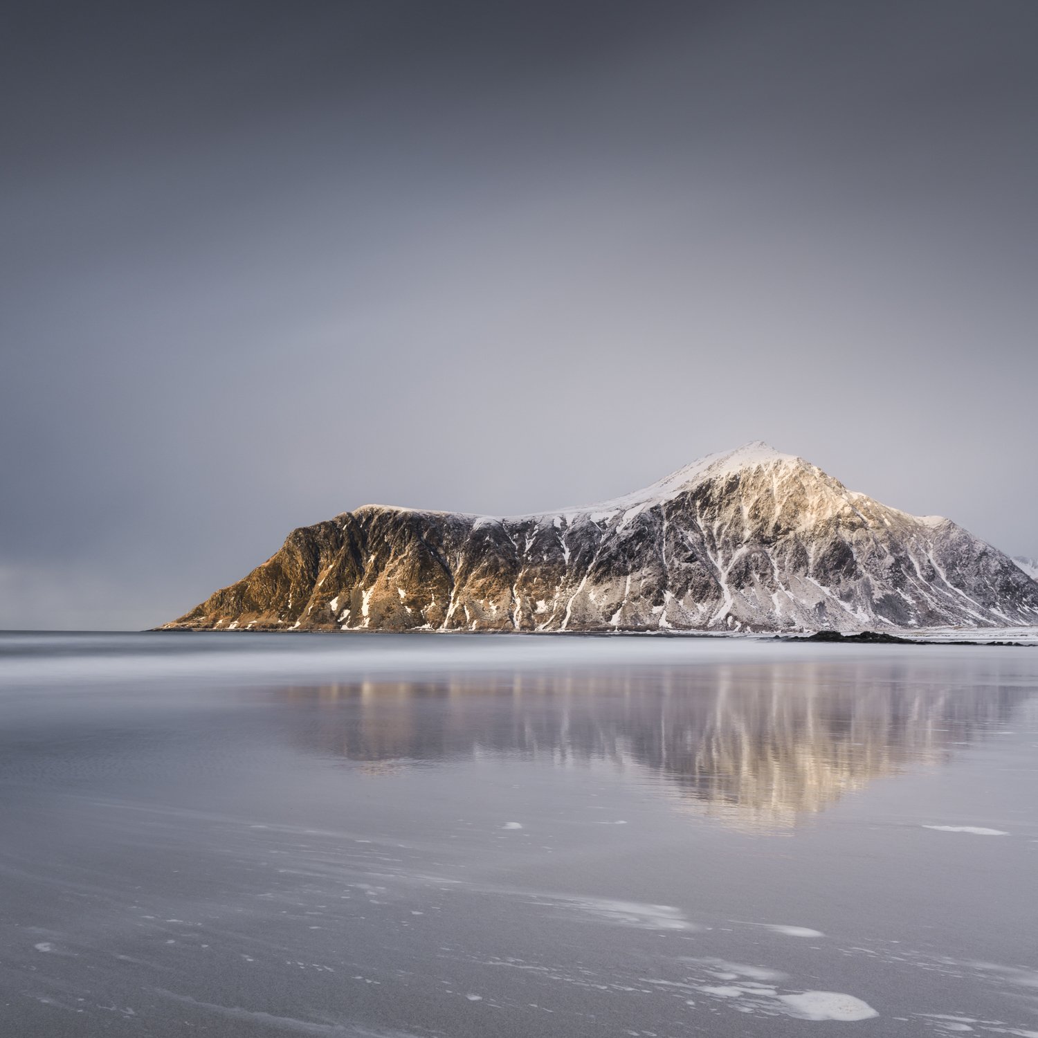 Reflections. Long-exposure photograph of a snow-covered mountain in Lofoten reflected in exceptionally calm water beneath a pale blue-grey sky.