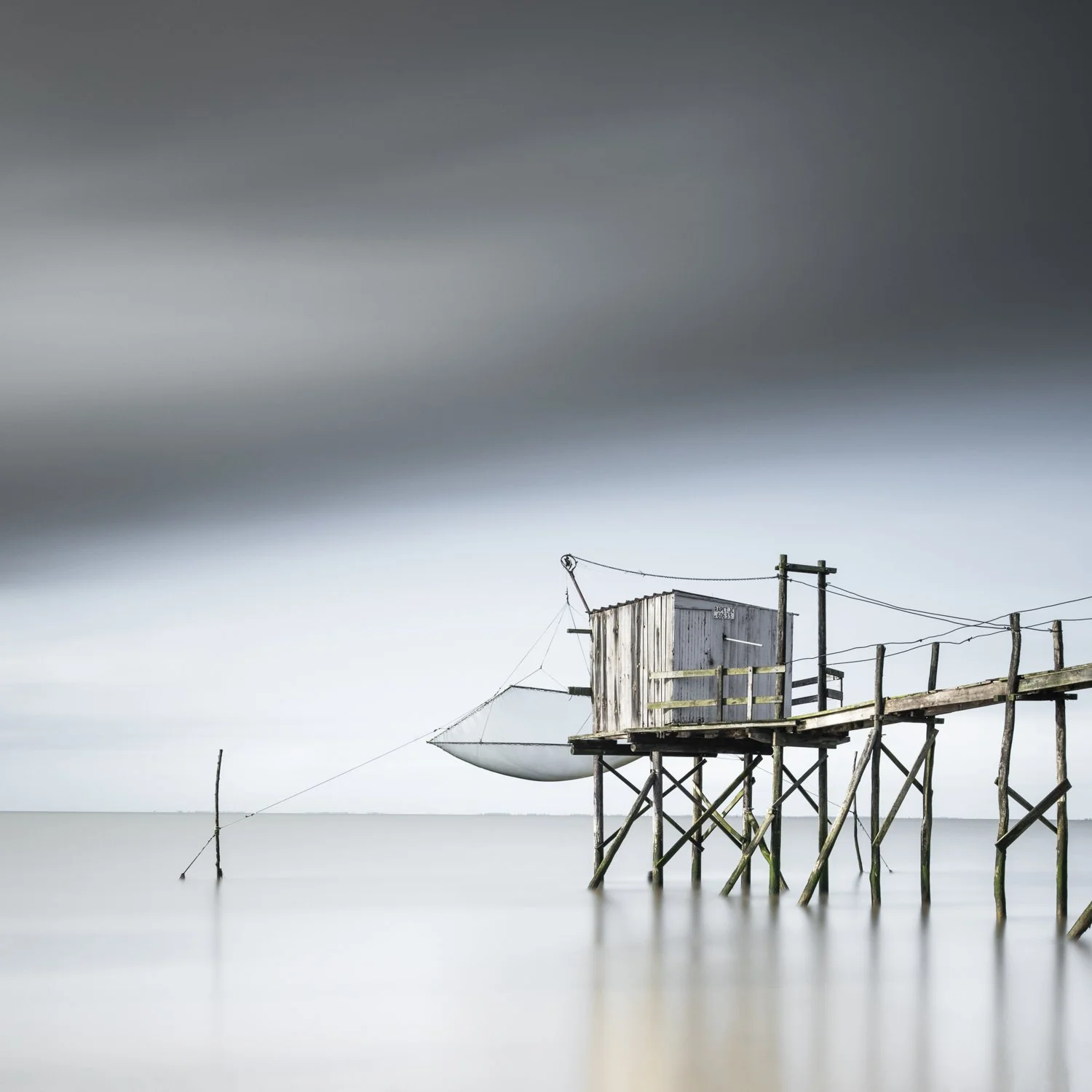 Fishing Hut II. Long-exposure photograph of a wooden fishing hut on stilts beside calm coastal water on the Gironde Estuary, with its reflection softened by the tide.
