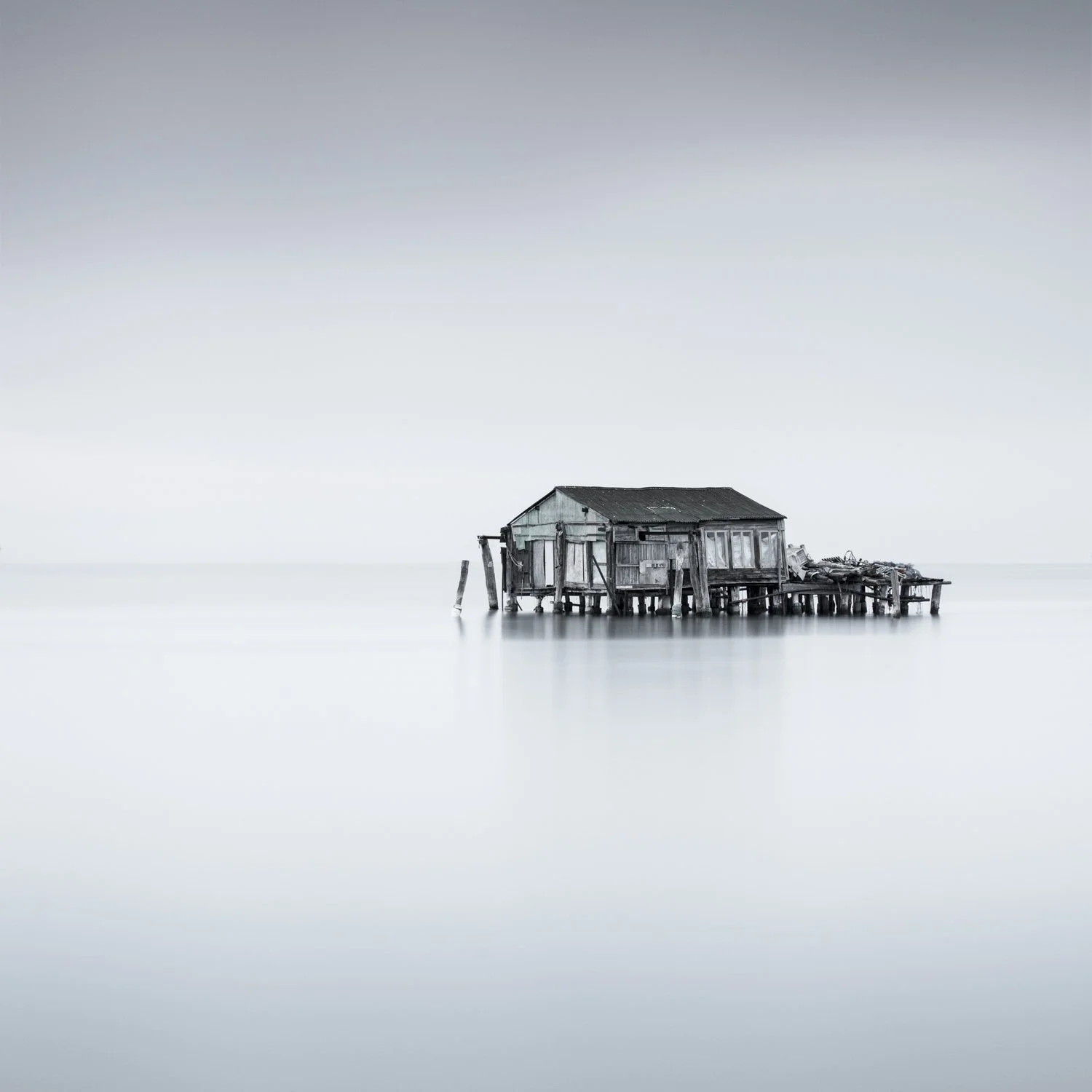 Alone. Long-exposure photograph of an isolated wooden fishing hut in the Venetian Lagoon at Pelistrino,  standing above calm water beneath a broad, pale sky.