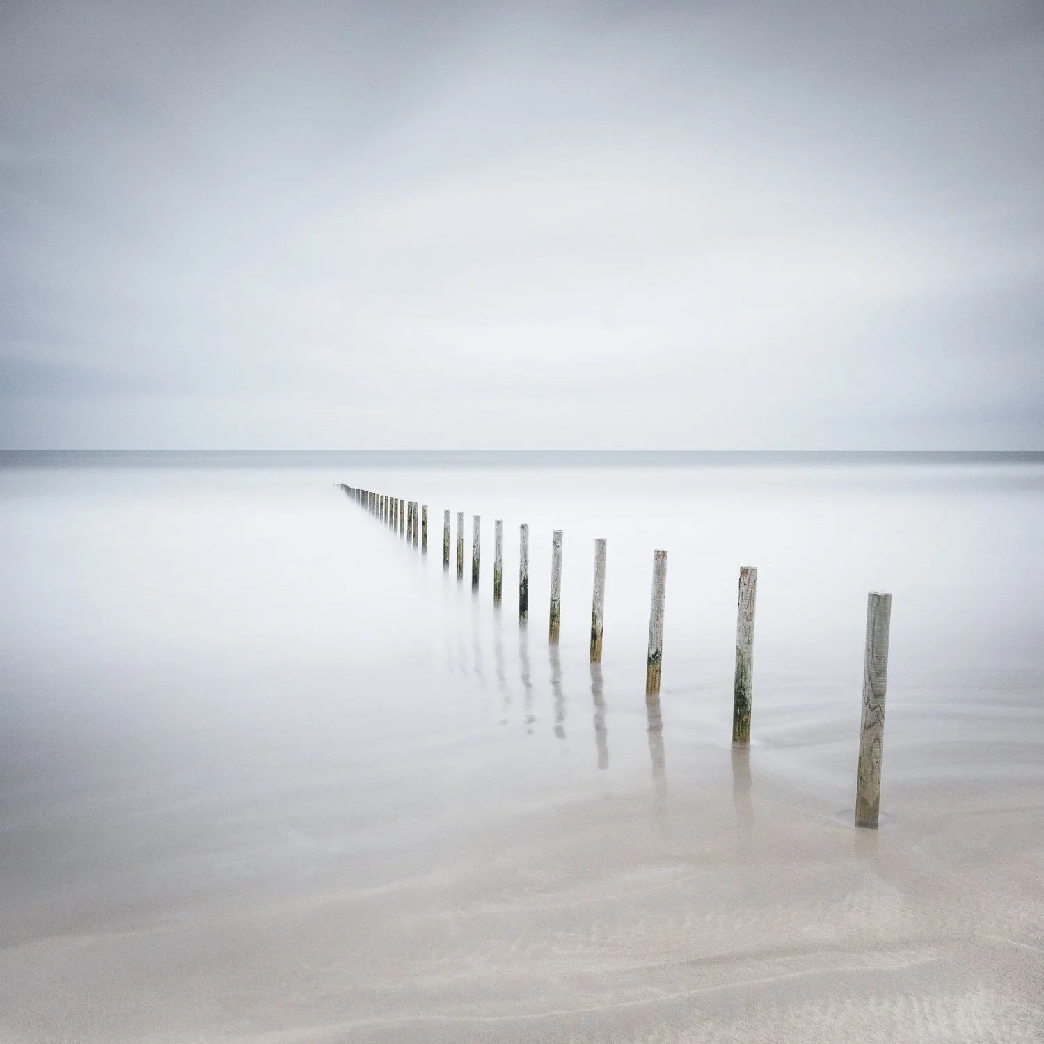 Passing Lines. Long-exposure photograph of a row of wooden posts receding into calm shallow water beneath a pale, misty sky on Portstewart Strand.