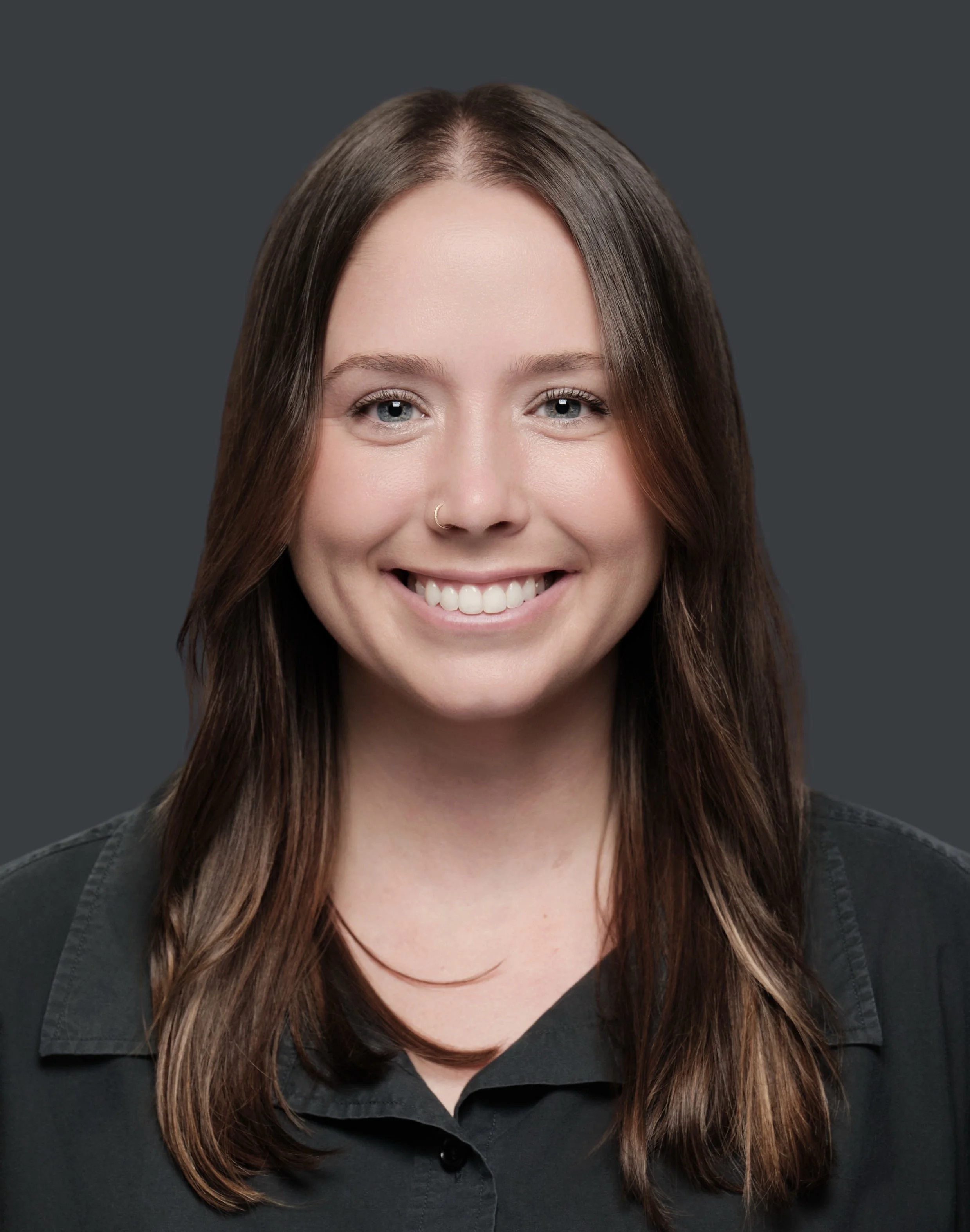 Close-up of a woman with long brown hair, blue eyes, a nose ring, smiling, wearing a black shirt, against a dark gray background.