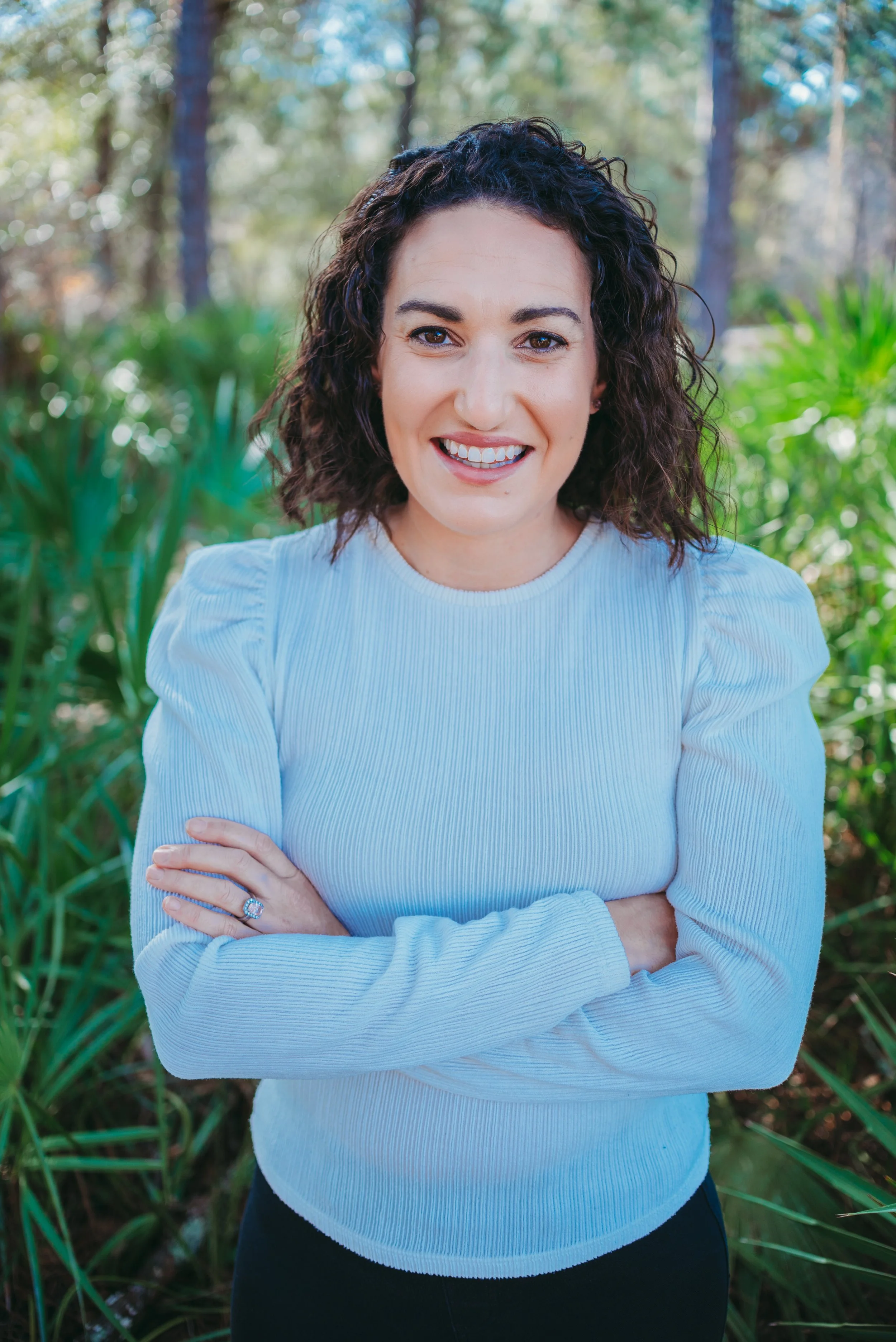 A woman with dark curly hair and a light blue long-sleeve shirt smiling outdoors in a lush, green forest setting.