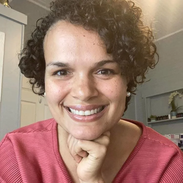 A woman with short curly hair, wearing a red top, smiling and resting her chin on her hand inside a room with light-colored walls and plants in the background.