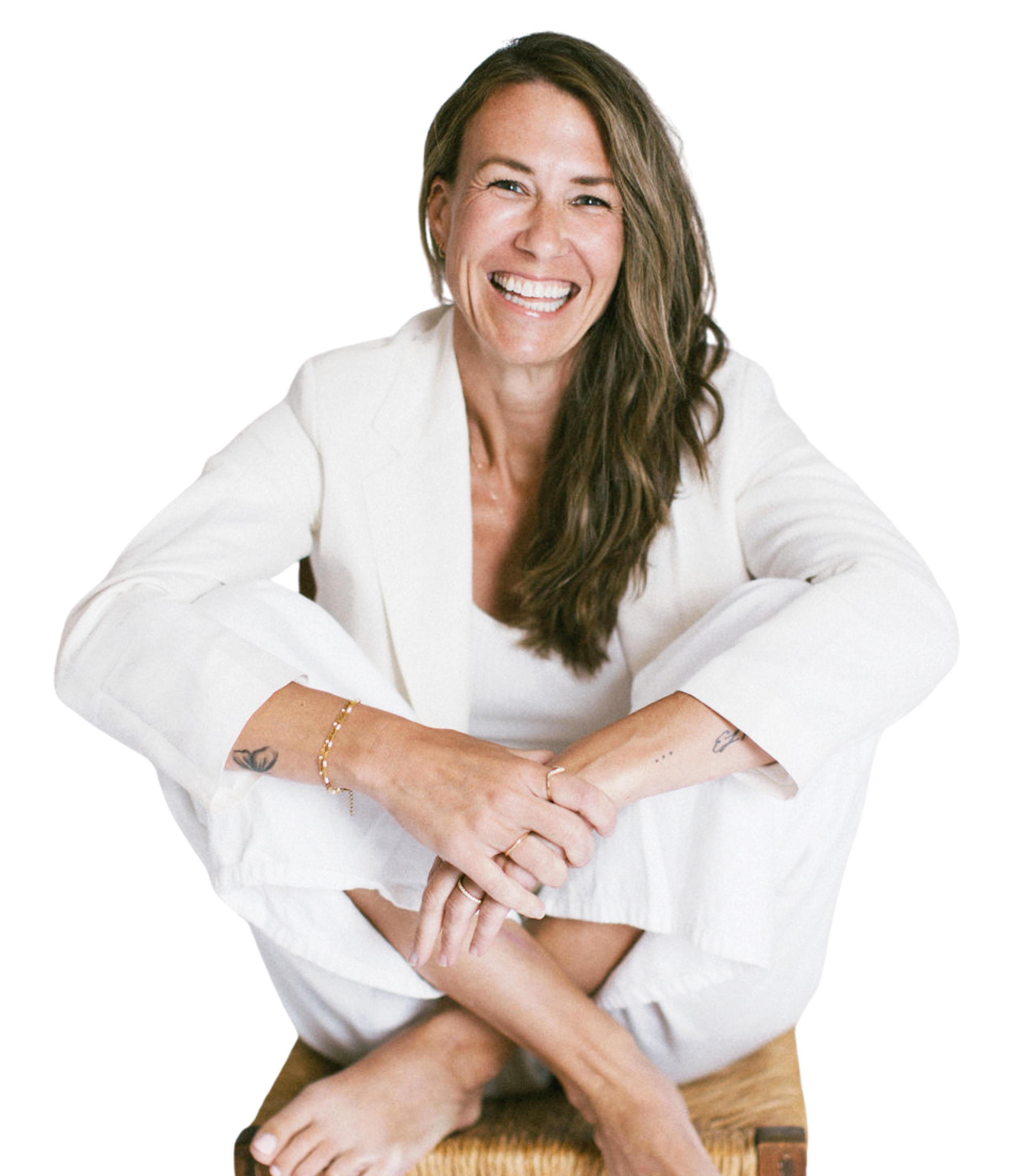 A woman with long brown hair and a white blazer smiling widely, sitting on a wooden floor, with her arms crossed and hands resting on her knees.