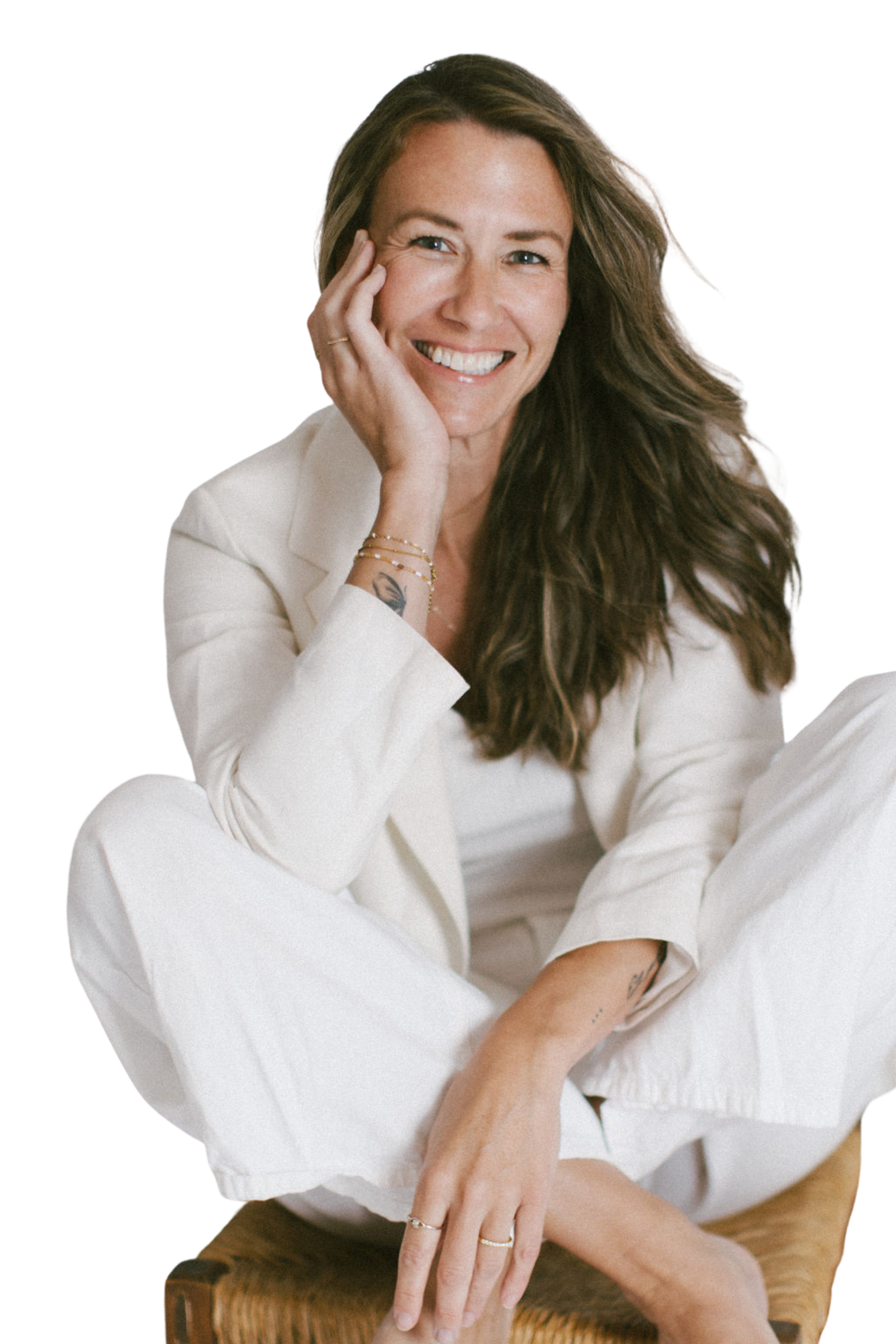 A woman with long brown hair, smiling, wearing an off-white blazer and pants, sitting on a wicker chair, with her hand resting on her face, displaying tattoos and jewelry.