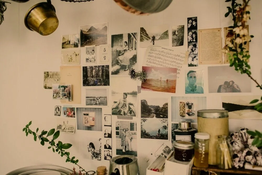 A wall decorated with various black-and-white and color photographs, notes, and artwork, with some greenery and a few metal containers and jars in the foreground.