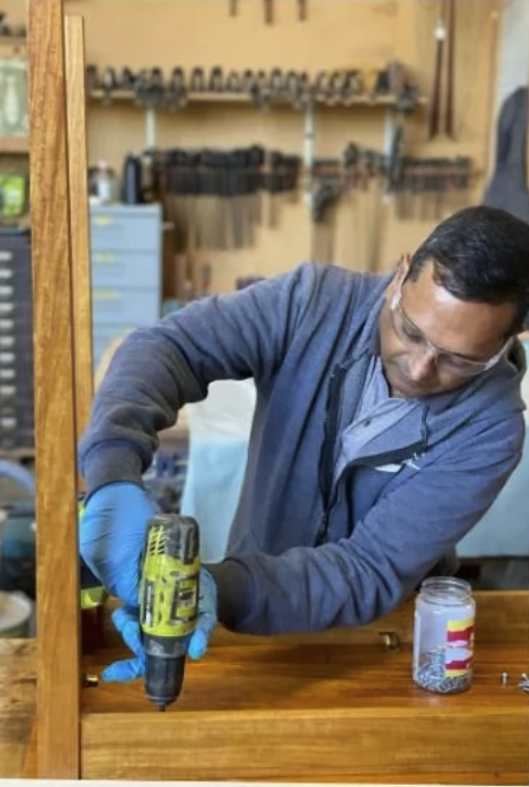 A man wearing safety glasses and blue gloves using a power drill to work on a wooden surface in a workshop.