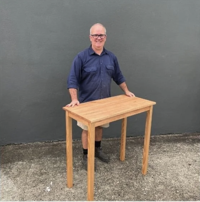 A man standing behind a wooden table outdoors against a gray wall, smiling at the camera.