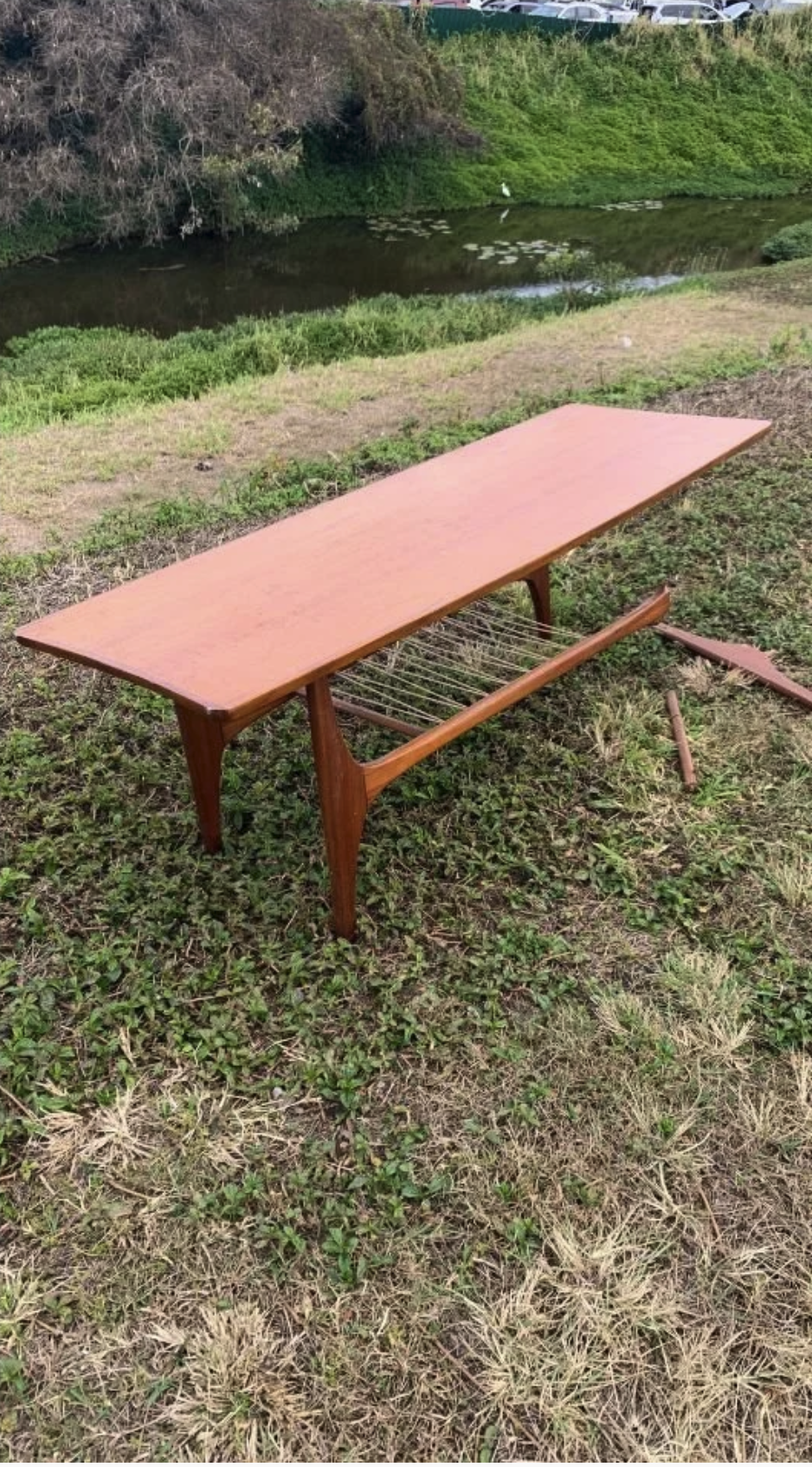 Disassembled wooden table with a glass or plastic top, placed on grass near a body of water with lily pads. The table parts are on the ground nearby.
