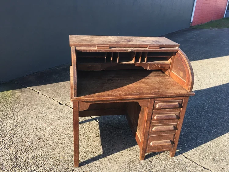 A vintage wooden roll-top desk with multiple drawers and compartments, situated outdoors on a concrete surface with a shadow cast on the ground.