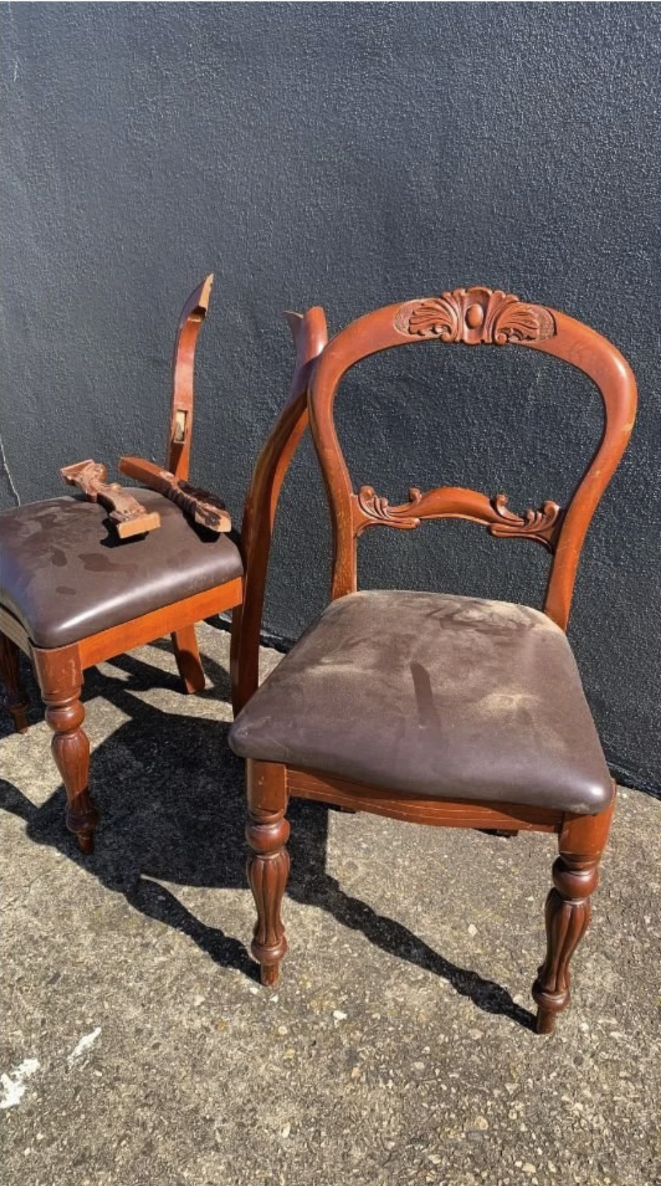 Two wooden chairs with carved backrests and upholstered seats are outdoors on a concrete surface against a dark wall. The chair in the foreground has a broken leg, with the upper part of the leg lying on the seat. The sunlight creates shadows of the 