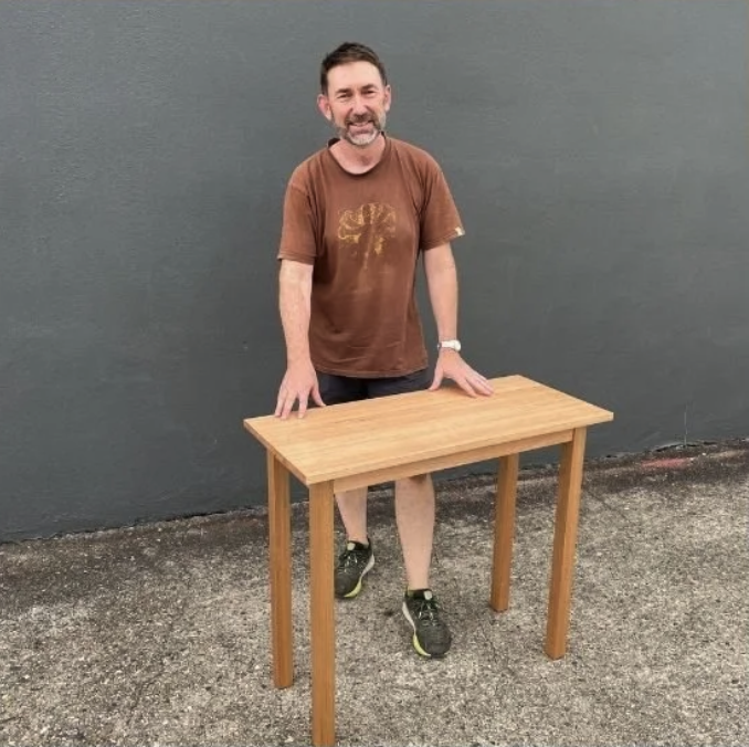 A man with gray hair and beard, wearing a brown t-shirt, black shorts, and black sneakers, is standing outdoors behind a small wooden table against a gray wall. His right hand is resting on the table.