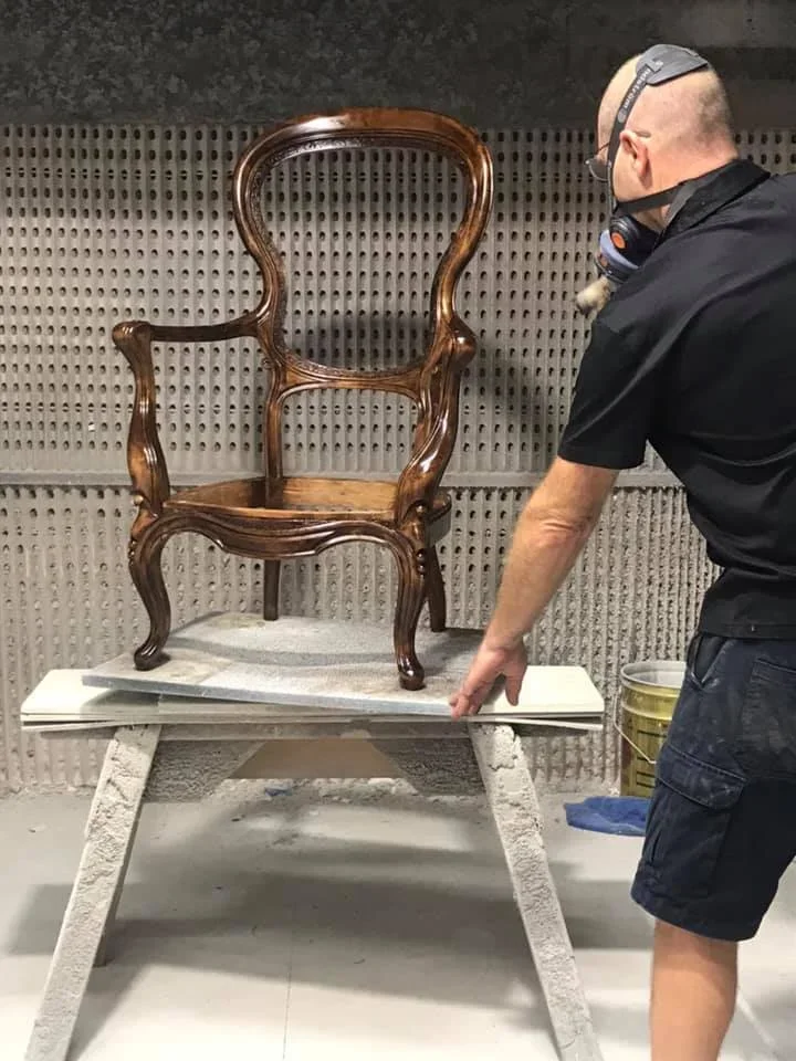 A person wearing a black shirt, shorts, safety goggles, and a mask points at a carved wooden chair placed on a white table inside a workshop with a textured wall.