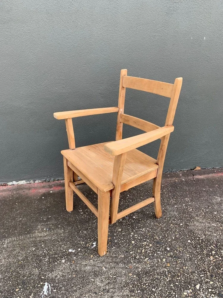 A wooden chair with armrests and a backrest, placed on a gravel surface against a gray wall.