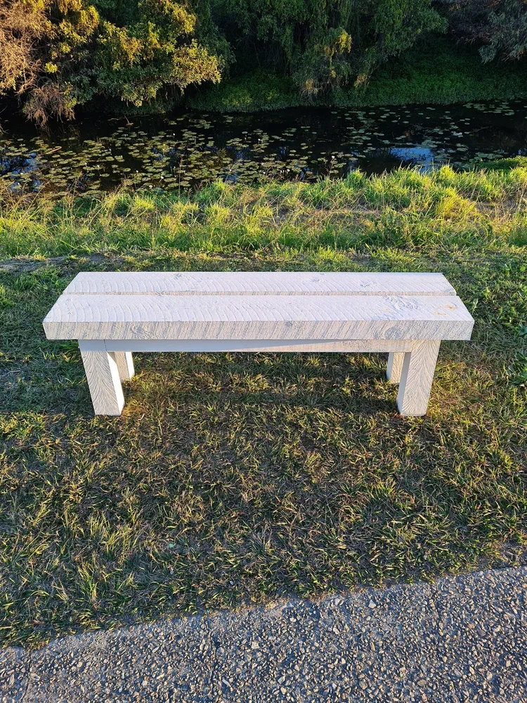 White wooden park bench on grassy ground near a pond with lily pads and green trees in the background.