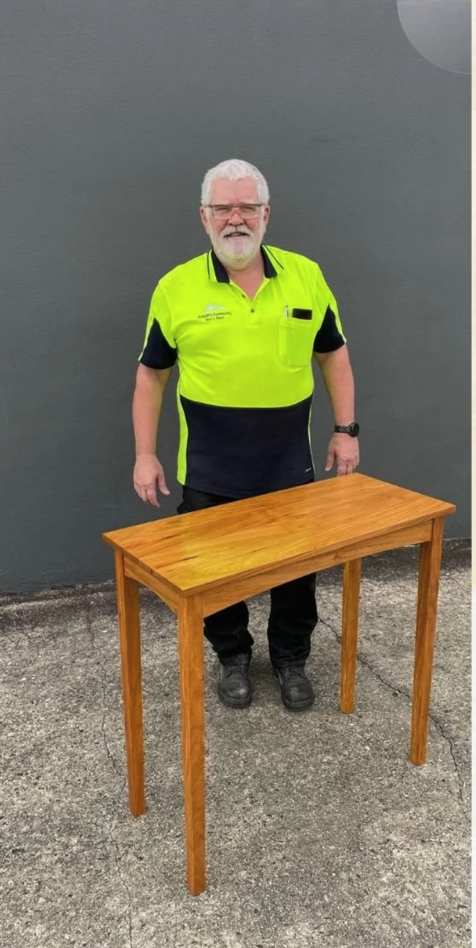 A man with white hair and a beard, wearing glasses and a high-visibility yellow and black shirt, standing outdoors next to a wooden table on a concrete surface against a gray wall.