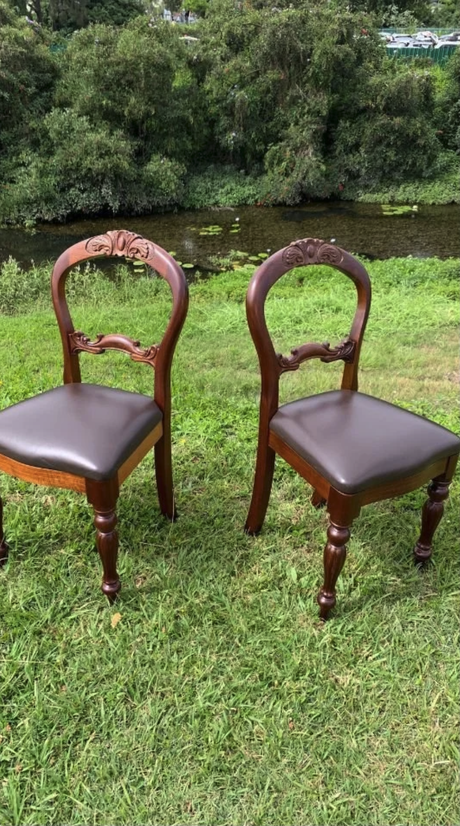 Two wooden chairs with upholstered seats are placed outside on green grass near a small pond with lily pads and surrounded by trees.