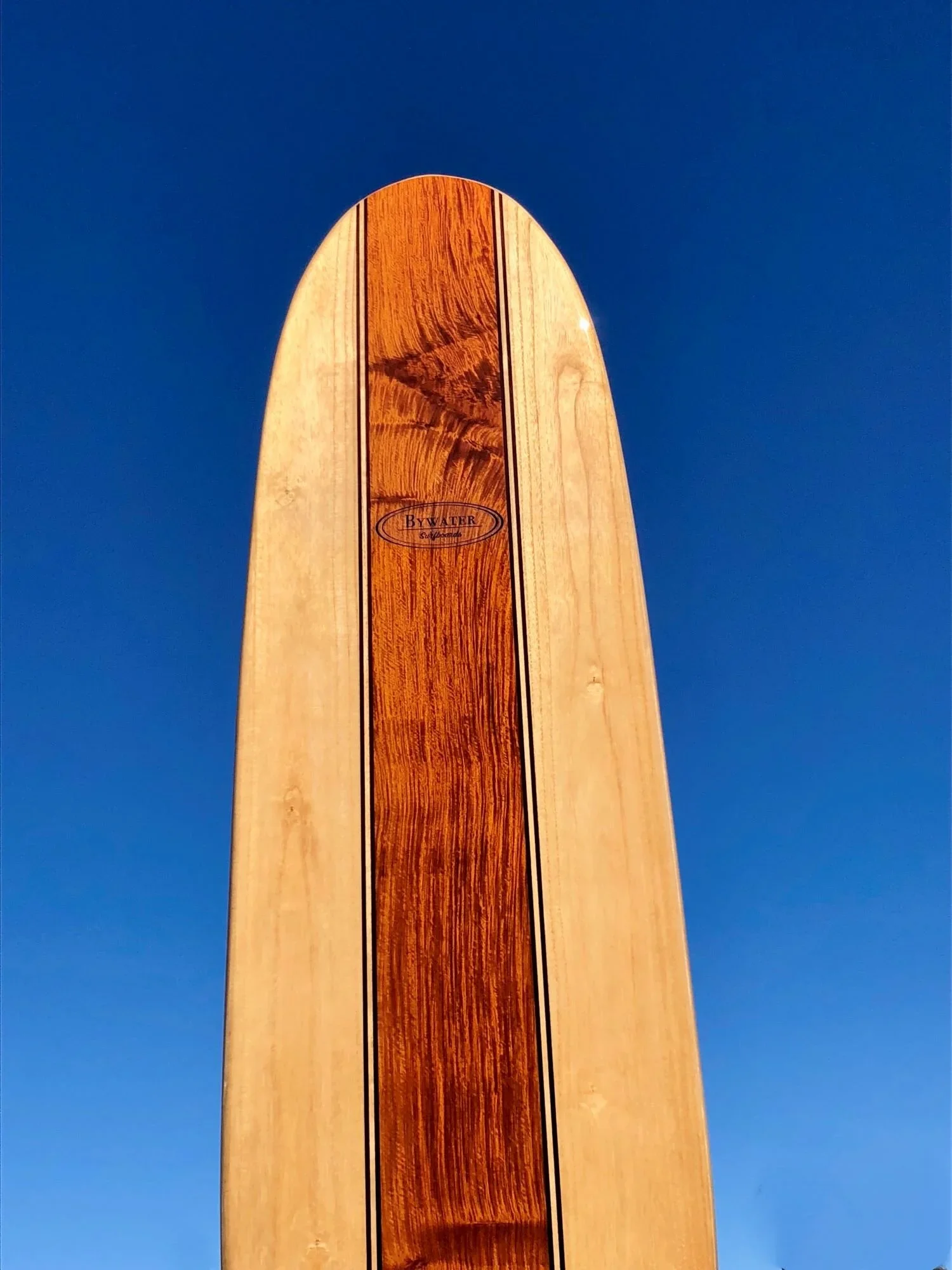 Close-up of a wooden surfboard with a logo reading "By Water Surfboards", held up against a clear blue sky.