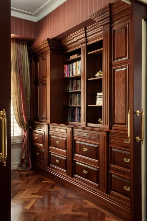 A large wooden built-in bookshelf and cabinet with brass handles in a room with herringbone hardwood floors and a window with velvet curtains.