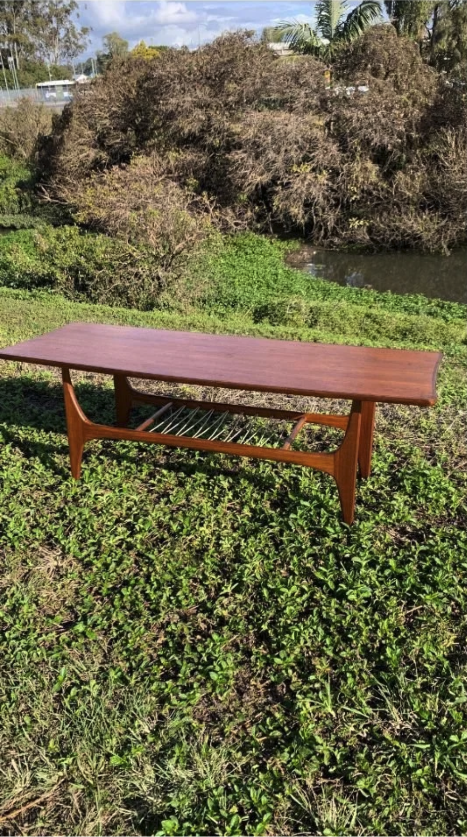 A wooden bench with a slanted seat and a lower rack for drying or storage, situated outdoors on green grass with bushes and trees in the background.