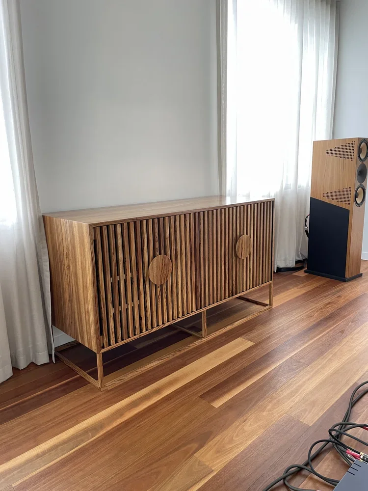 A wooden sideboard with slatted doors next to a large modern speaker in a room with wood flooring and white curtains.
