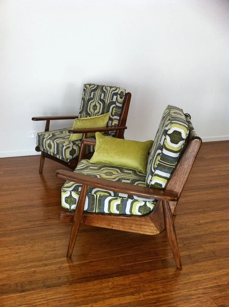 Two mid-century modern armchairs with wooden frames and patterned upholstery, each topped with a green velvet pillow, positioned on a wooden floor against a plain white wall.