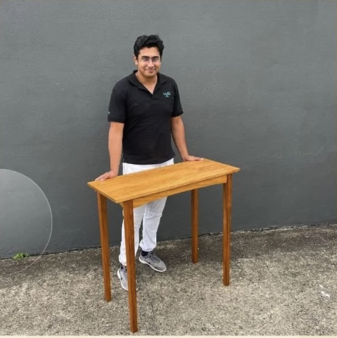 A man in a black shirt and white pants standing outdoors next to a small wooden table with a gray wall in the background.