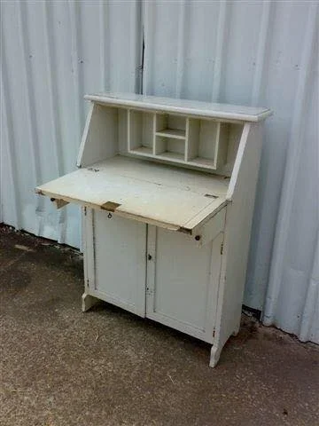 White wooden secretary desk with small upper shelves and closed lower cabinet, placed outdoors against a corrugated metal wall.