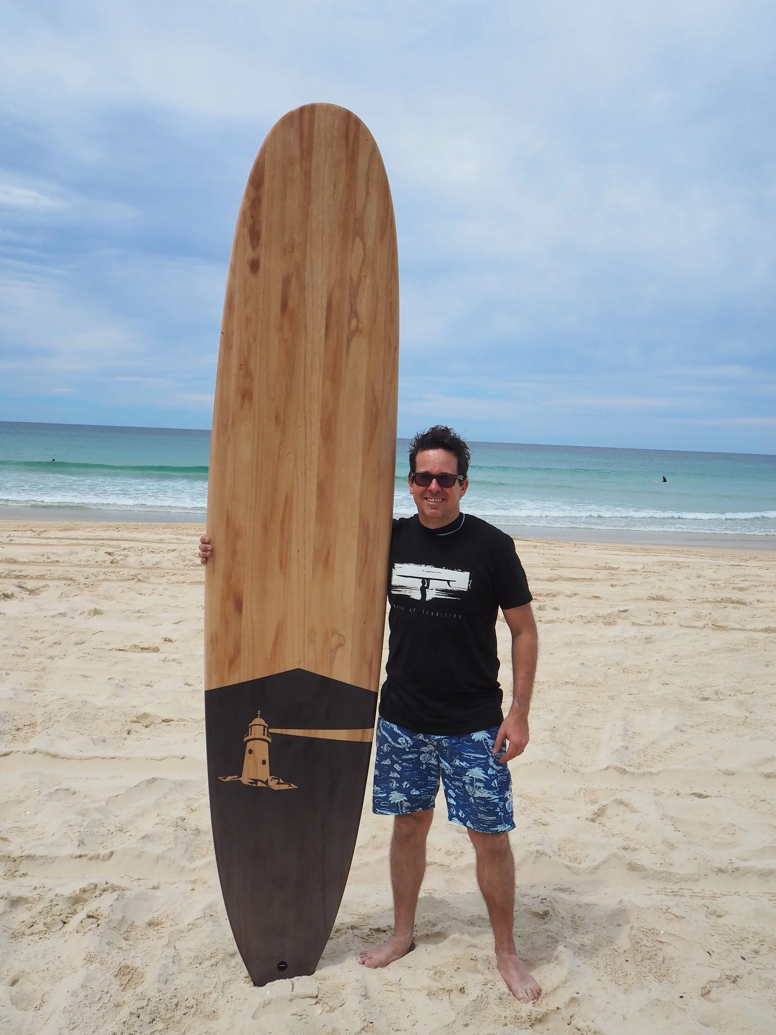 A man standing on a sandy beach holding a large wooden surfboard with a lighthouse illustration at the bottom. The man is wearing sunglasses, a black T-shirt, and blue patterned swim shorts, smiling, with the ocean and cloudy sky in the background.