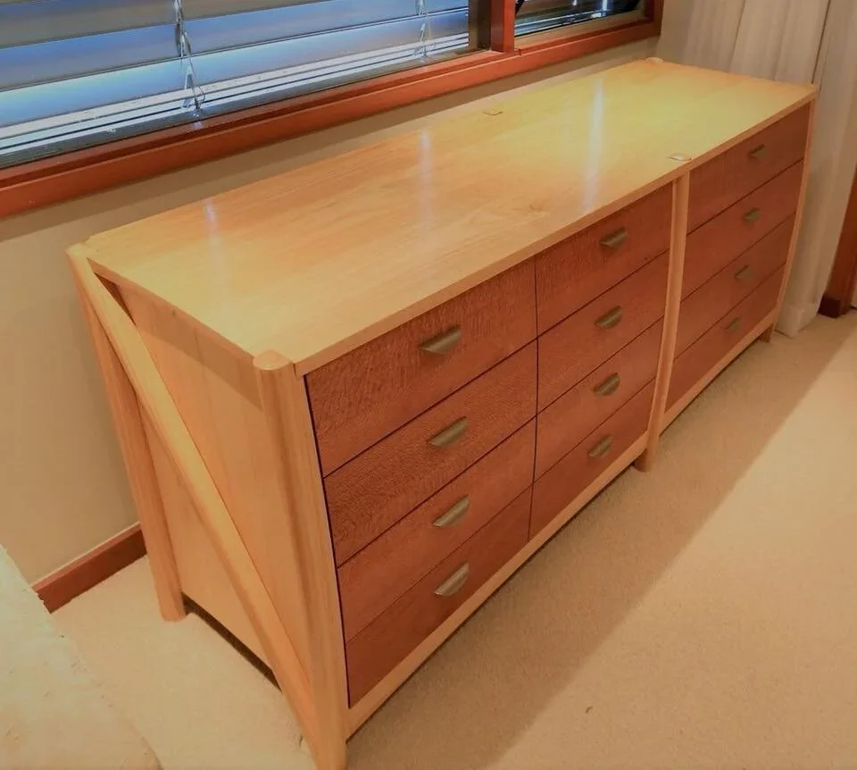 A wooden dresser with horizontal drawers and brass handles, positioned underneath a window with partially opened blinds.