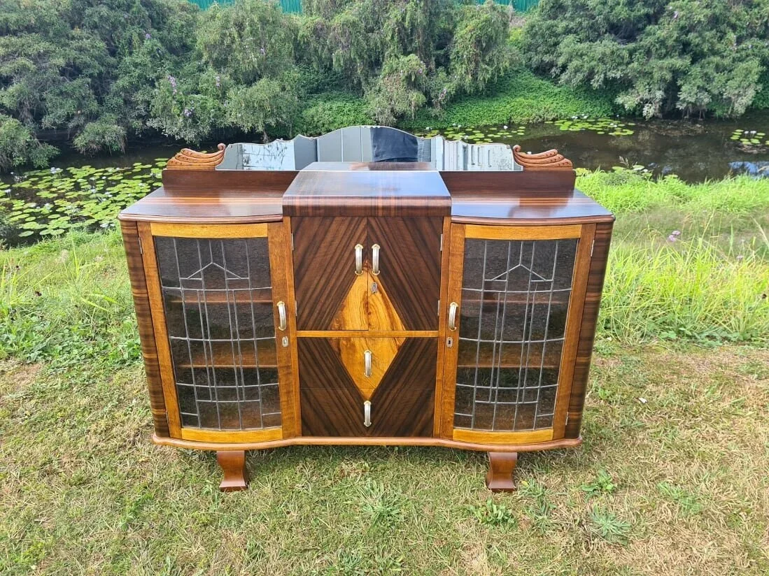 A vintage wooden cabinet with glass doors and decorative carvings, placed outdoors on grass near a pond with lily pads and trees in the background.