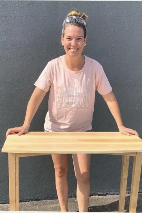 Woman with glasses on her head smiling, standing outdoors behind a wooden table against a gray wall, and holding onto it with both hands.