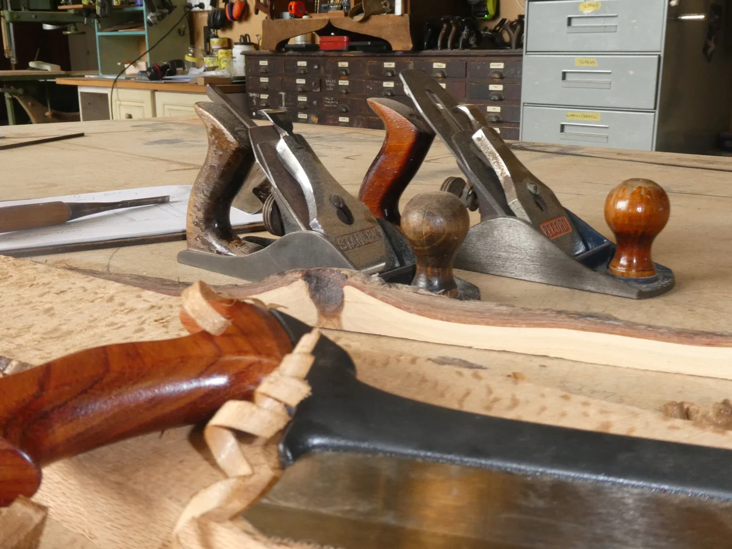 A woodworking workbench with hand planes, a carving knife, and a piece of wood in the workshop.