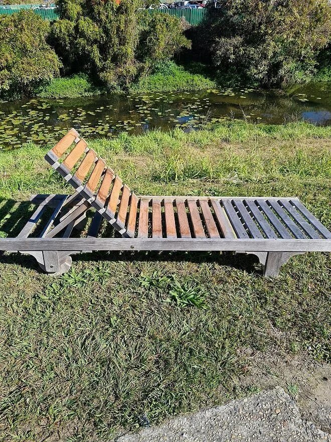 A wooden lounge chair with adjustable backrest, placed on green grass near a pond with lily pads, surrounded by trees and greenery.