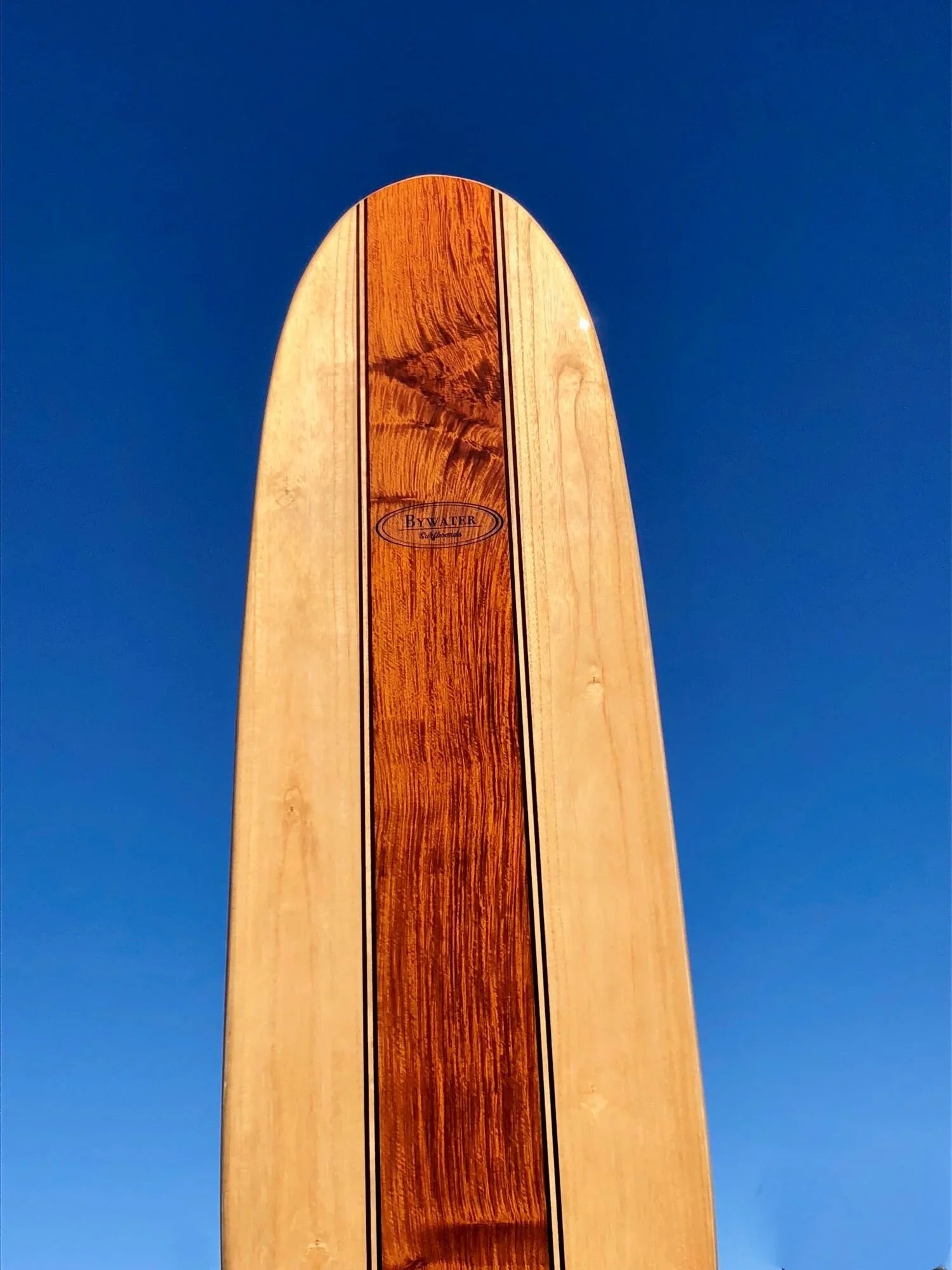 Close-up of a wooden surfboard with a logo, set against a clear blue sky.