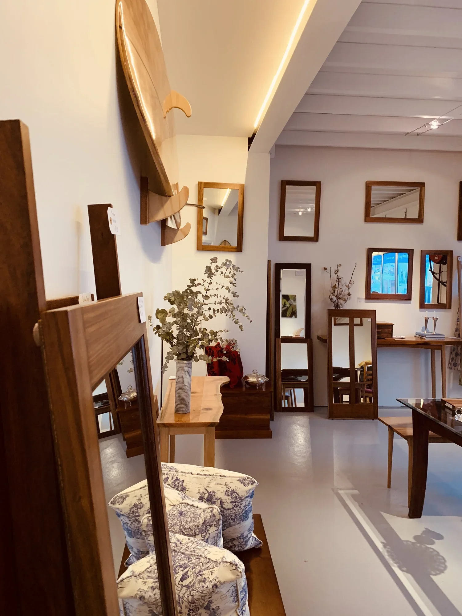 Interior of a furniture store displaying wooden tables, chairs, mirrors, and a sofa with floral upholstery, with sunlight casting shadows through a window.