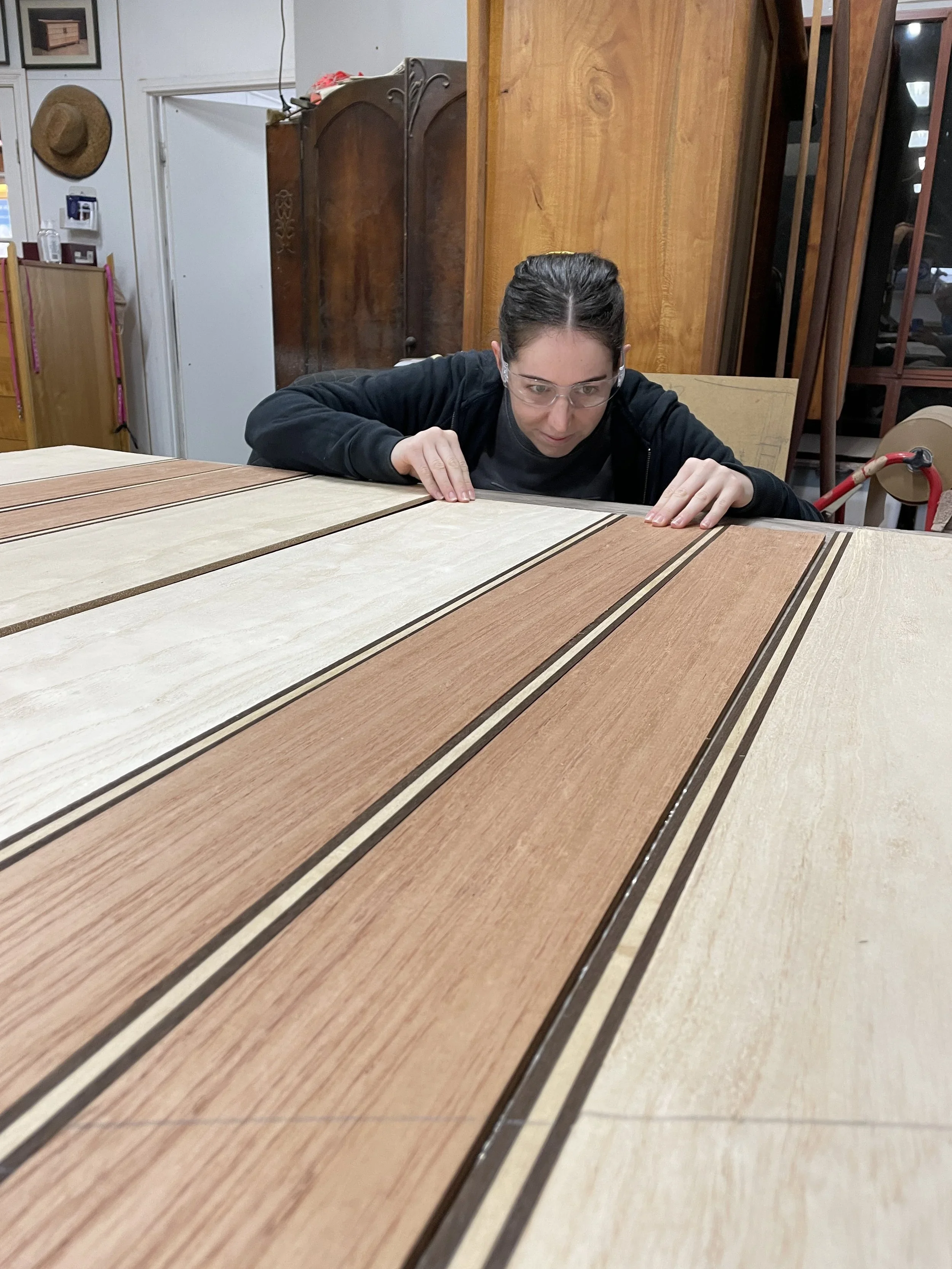 A woman inspecting a wooden table in a workshop or furniture store, with wood panels and furniture in the background.