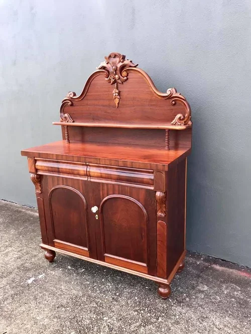 A vintage wooden cabinet with an ornate carved top, standing against a gray wall on a concrete surface.