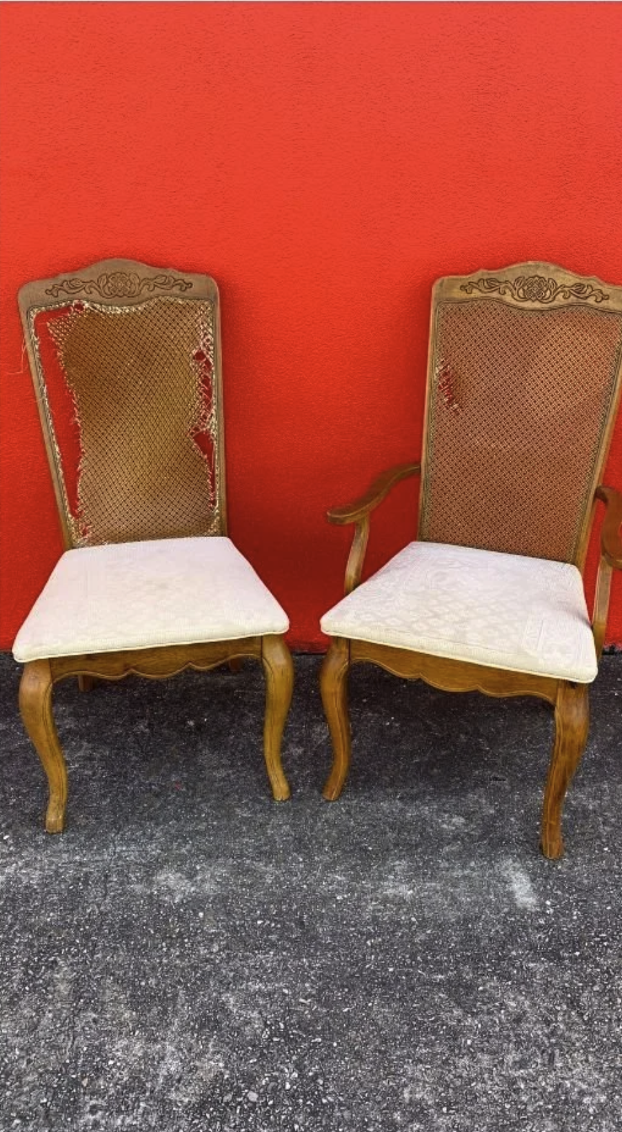 Two vintage wooden chairs with intricate carvings, woven backrests, and padded white seats, positioned against a bright red wall with a textured surface, on a dark concrete floor.